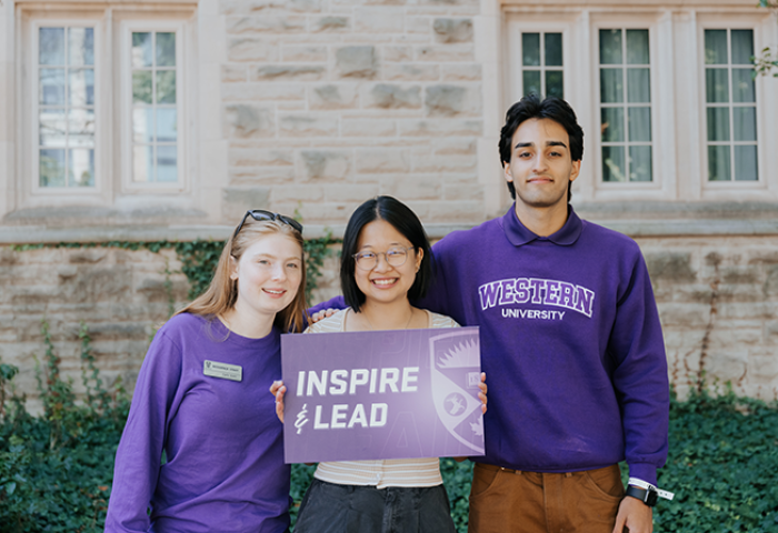 Three student leaders standing in front of residence holding a sign